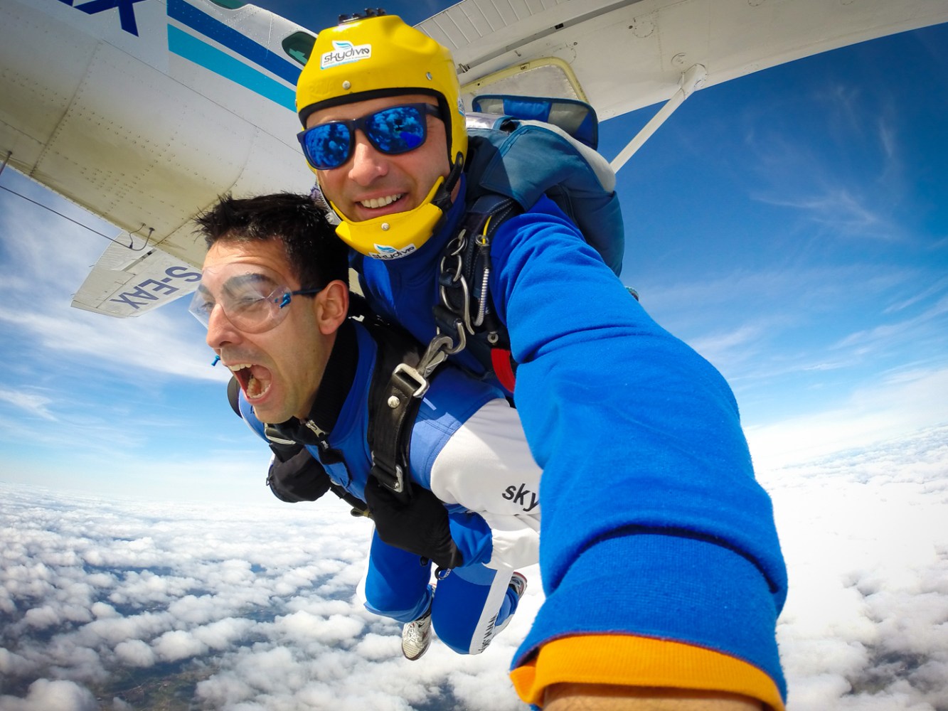 Two people tandem skydiving, smiling and excited, with clouds and sky in background.