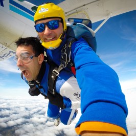 Two people tandem skydiving, smiling and excited, with clouds and sky in background.