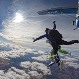 Two skydivers jump from a plane above clouds with sun shining brightly.