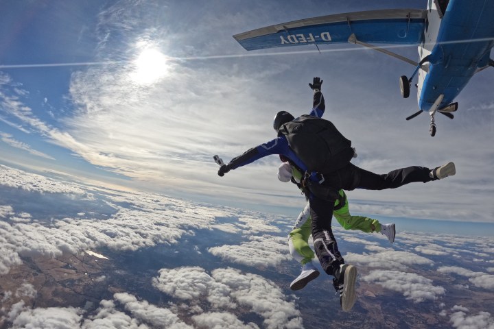 Two skydivers jump from a plane above clouds with sun shining brightly.