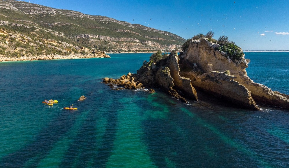 Kayakers near rocky outcrop in clear blue water with distant mountains and clear sky.