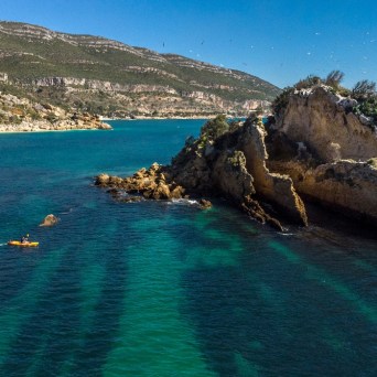 Kayakers near rocky outcrop in clear blue water with distant mountains and clear sky.