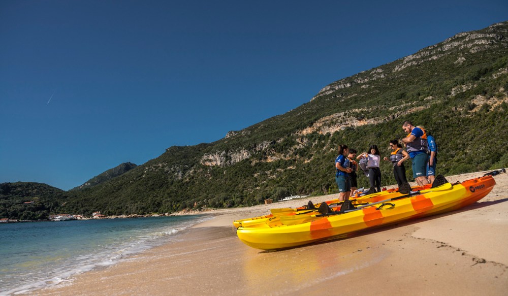 Group on beach with kayaks, rocky hills and clear blue sky in background.
