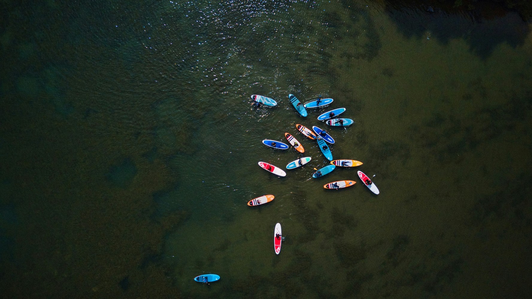 Aerial view of people on colorful paddleboards clustered on calm water.