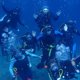 Eight scuba divers underwater, posing with raised hands and bubbles around them.