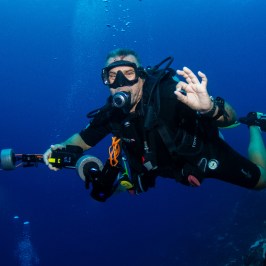 Scuba diver underwater holding a camera and gesturing OK sign with hand.