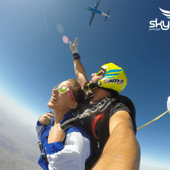 Tandem skydive with a parachute opening against a clear blue sky, airplane visible above.