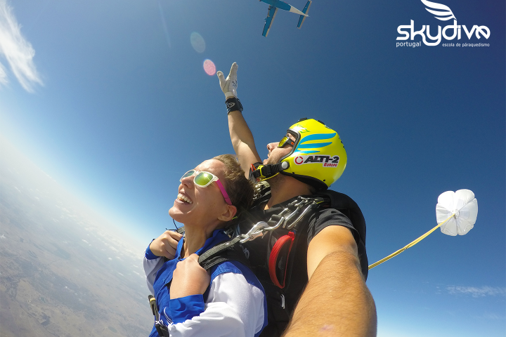 Tandem skydive with a parachute opening against a clear blue sky, airplane visible above.