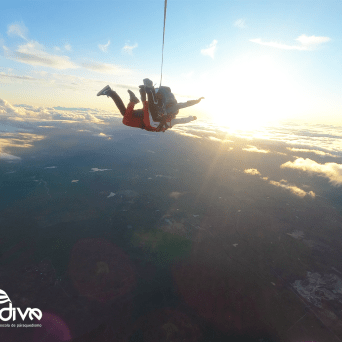 Two skydivers in tandem jump, silhouetted against a bright sunset above clouds.