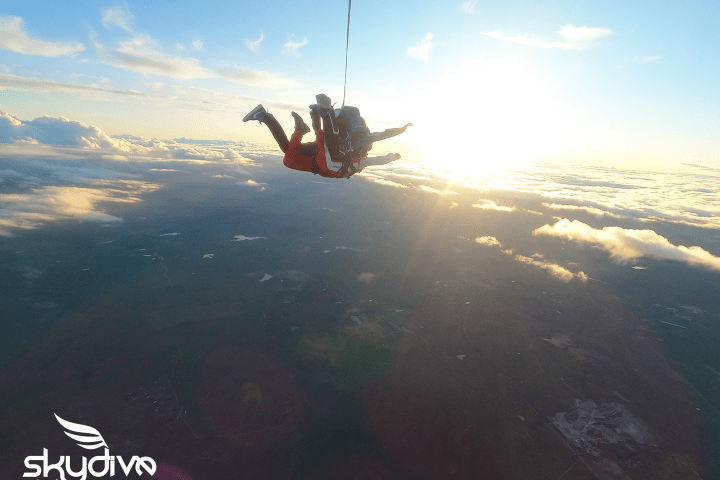Two skydivers in tandem jump, silhouetted against a bright sunset above clouds.