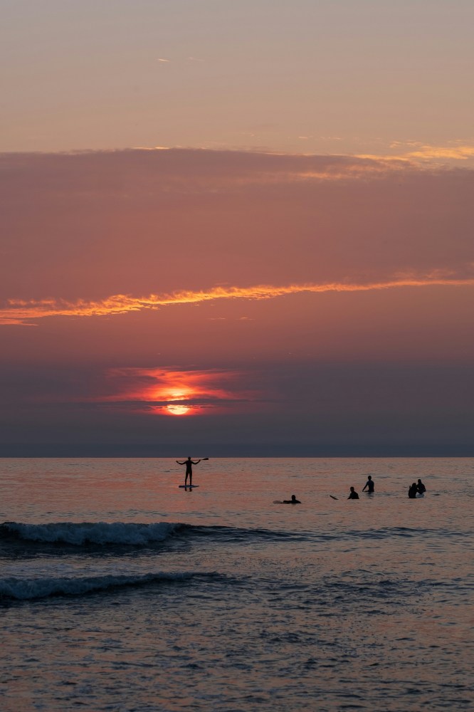Sunset over ocean with silhouette of paddleboarder and swimmers.
