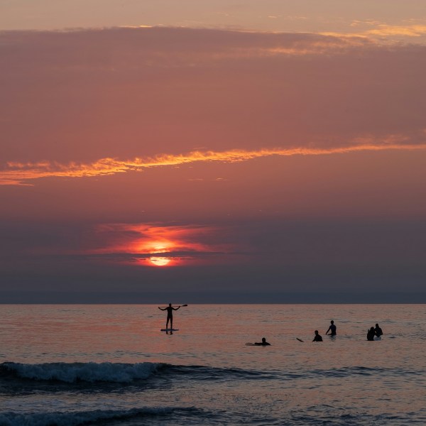 Sunset over ocean with silhouette of paddleboarder and swimmers.